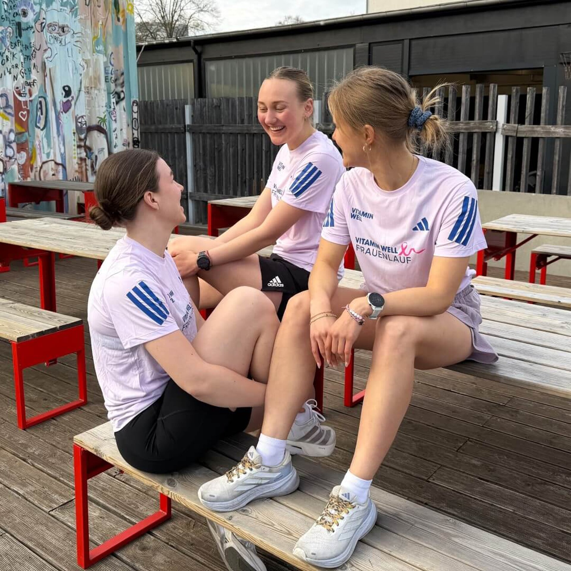 Drei Frauen im rosafarbenen Event-Shirt. Zentral auf der Brust das Frauenlauf-Logo mit pinker Brustkrebs-Schleife. Darüber links das adidas-Logo, rechts das VITAMIN WELL-Logo in Dunkelblau. adidas-Streifen auf den Ärmeln ebenfalls dunkelblau.