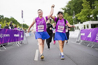 Zwei Frauen in T-Shirts des 40. VITAMIN WELL Frauenlauf Berlin laufen lächelnd Hand in Hand auf der Strecke.